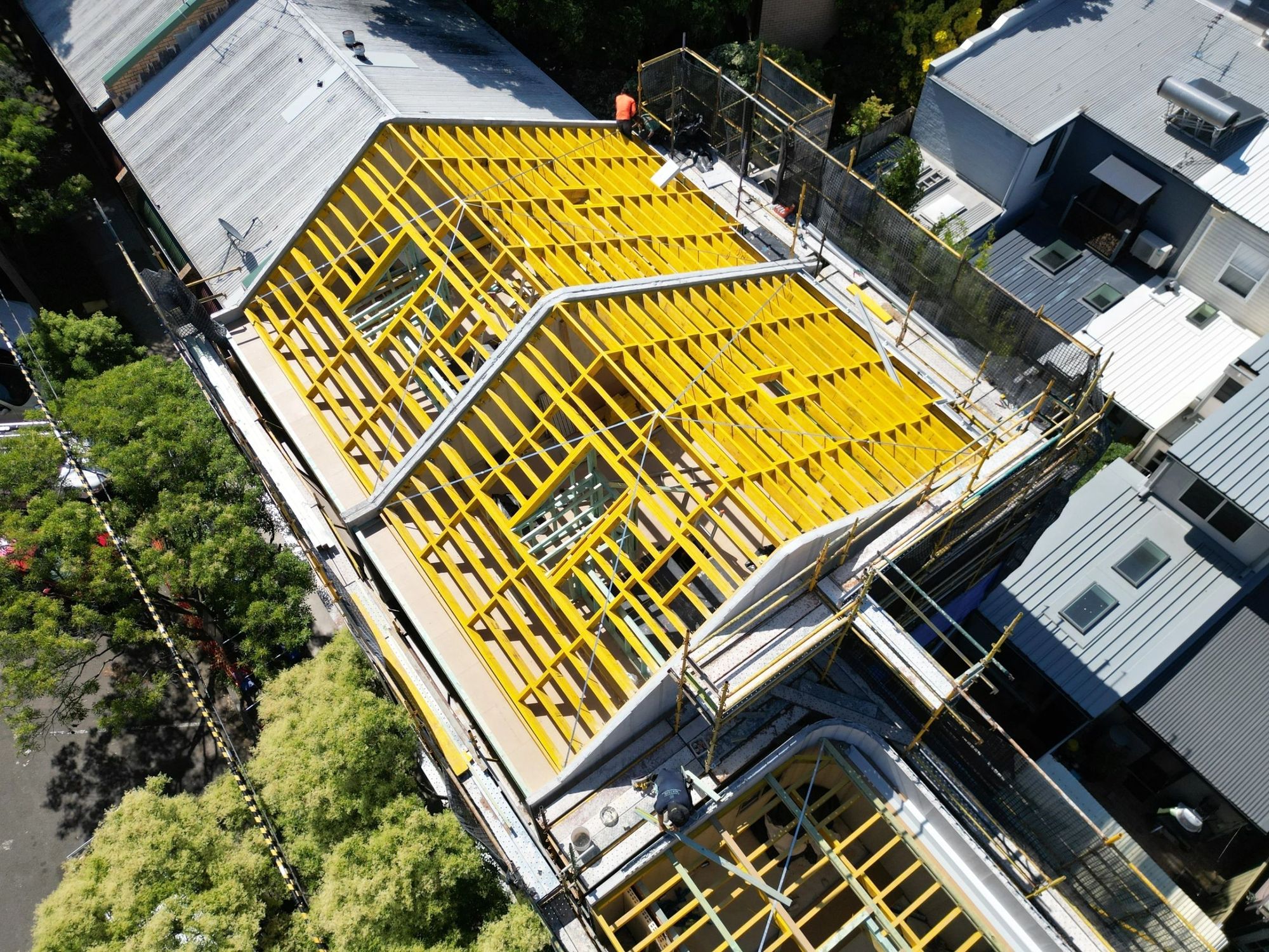 Aerial view of a completed timber roof frame, rafters and hips picked out in yellow, scaffolding around the perimeter of an inner-city addition.